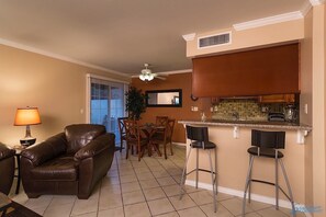 A living room with a brown leather sofa, a dining table with chairs, and a kitchen area with a bar stool.