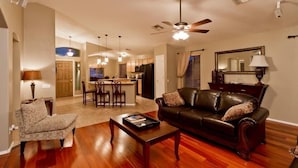 A spacious living room with a dark leather sofa, a patterned armchair, and a wooden coffee table. A ceiling fan and pendant lights are visible. - All Star By Signature Vacation Rentals (Chandler)