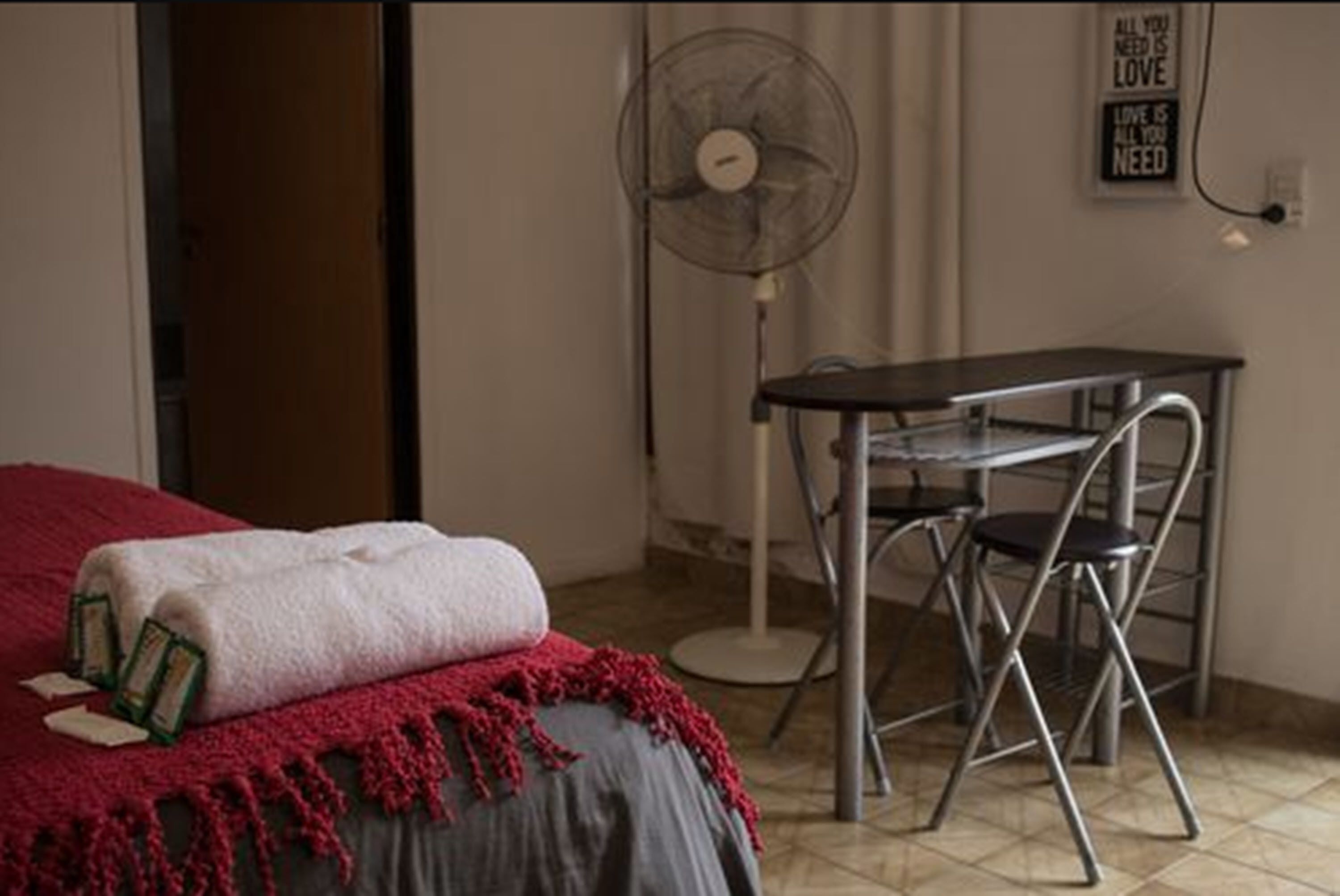 A bedroom with a bed, a small table with a chair, a standing fan, and a wall with a motivational quote.