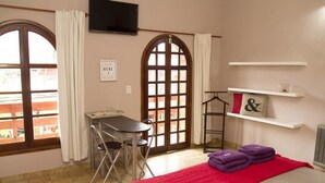 Una habitación con cama, escritorio y silla. Hay dos ventanas con cortinas, un televisor en la pared y una repisa con toallas.