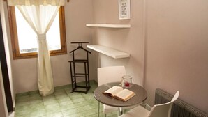 Una habitación pequeña con ventana, una mesa con un libro abierto, una silla, un radiador y una estantería con una cita motivacional.