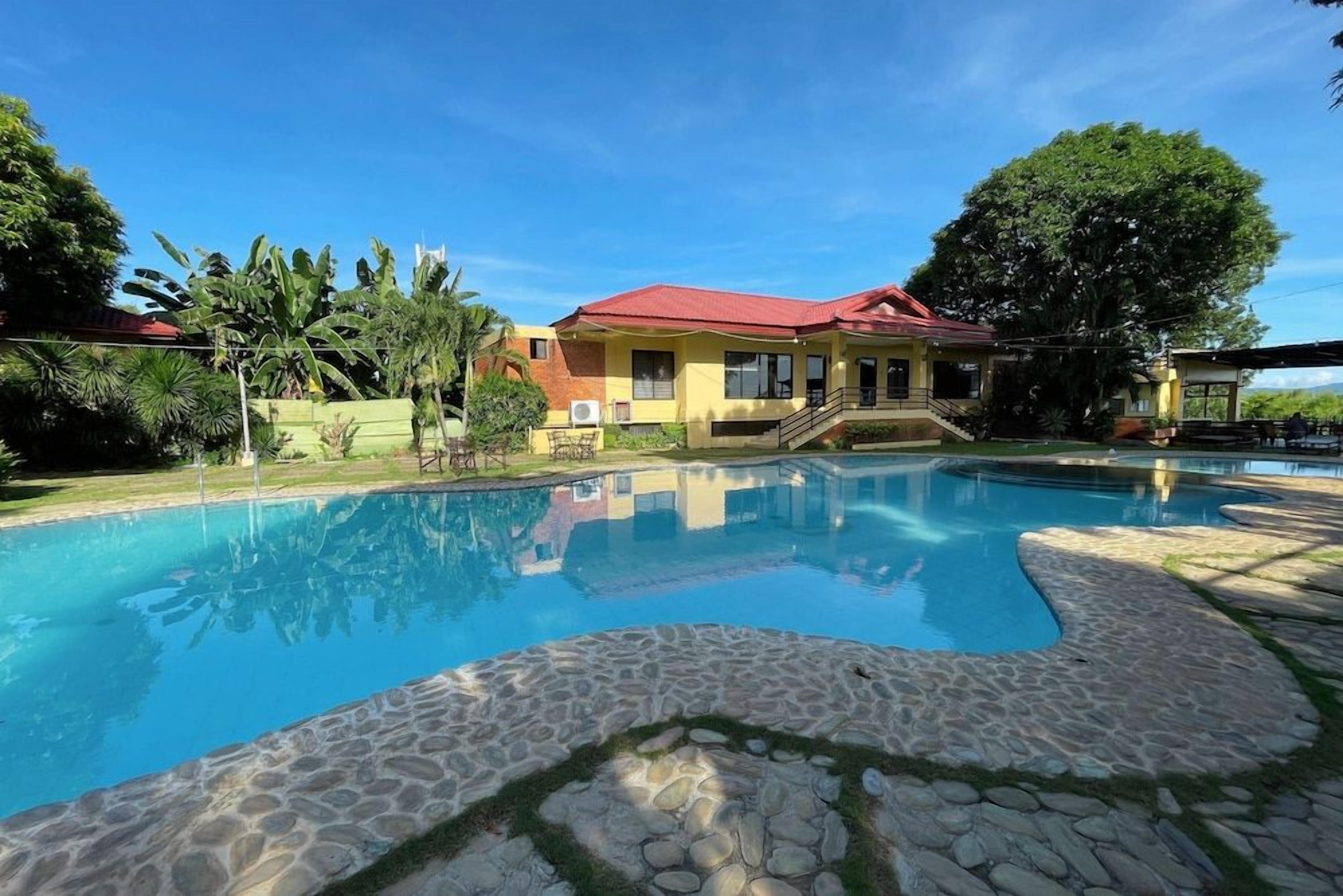 A swimming pool with a stone border, a building with a red roof, and lush greenery.