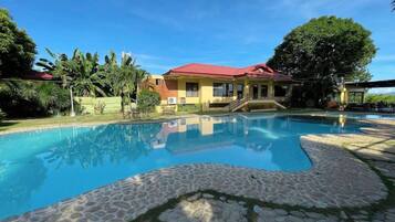 A swimming pool with a stone border, a building with a red roof, and lush greenery.