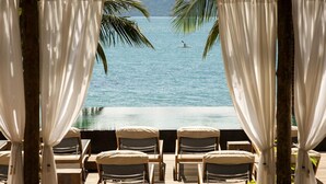 An infinity pool with lounge chairs and curtains, overlooking a calm sea with a person paddleboarding. - TW Guaimbê Exclusive Suites (Ilhabela)