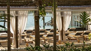 A sunlit outdoor lounge area with wooden loungers and white curtains, overlooking a pool and a wooden deck. - TW Guaimbê Exclusive Suites (Ilhabela)