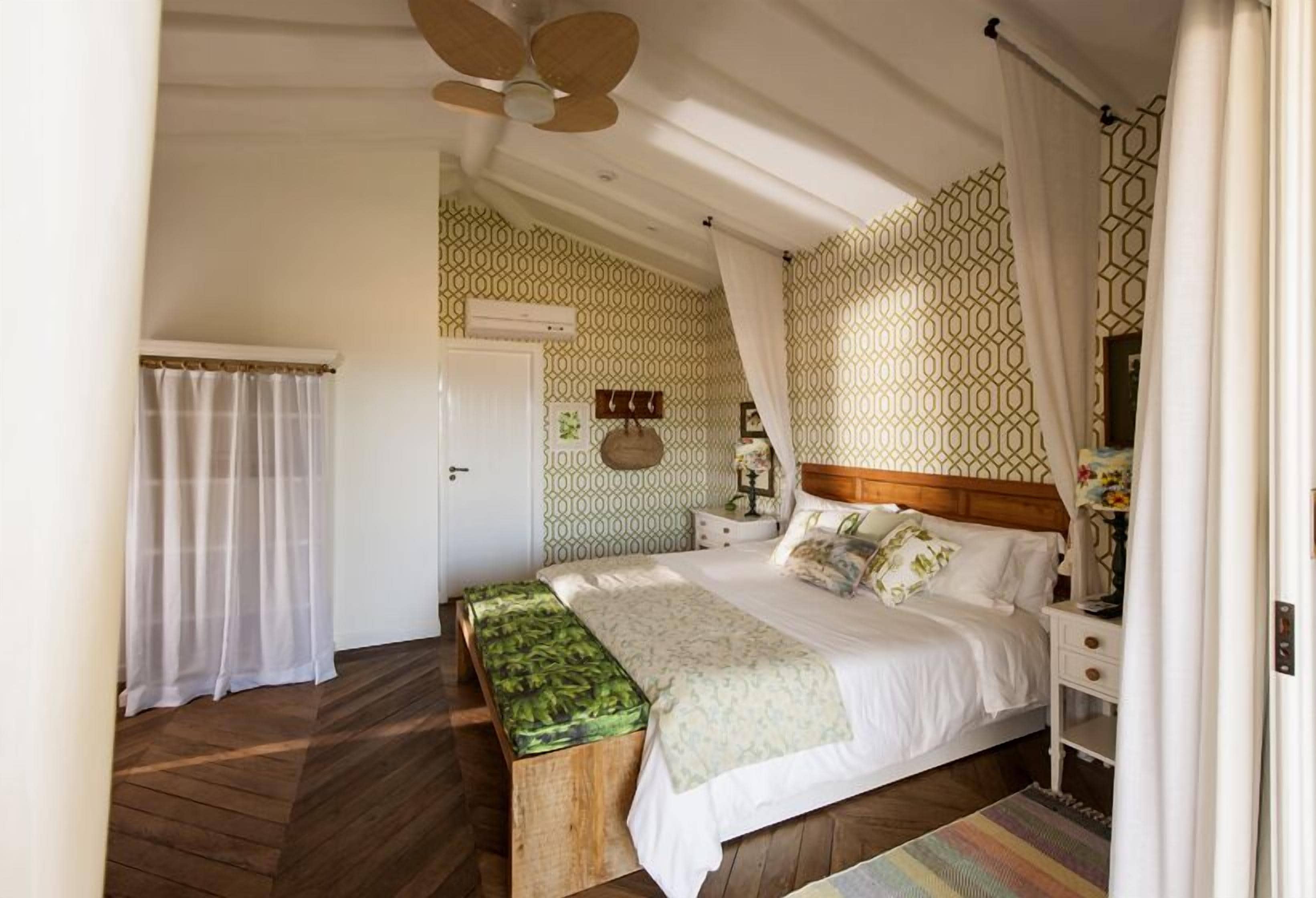 Un dormitorio con piso de madera, una cama con cubrecama blanco, un banco con funda verde, una mesita de noche y un ventilador de techo.