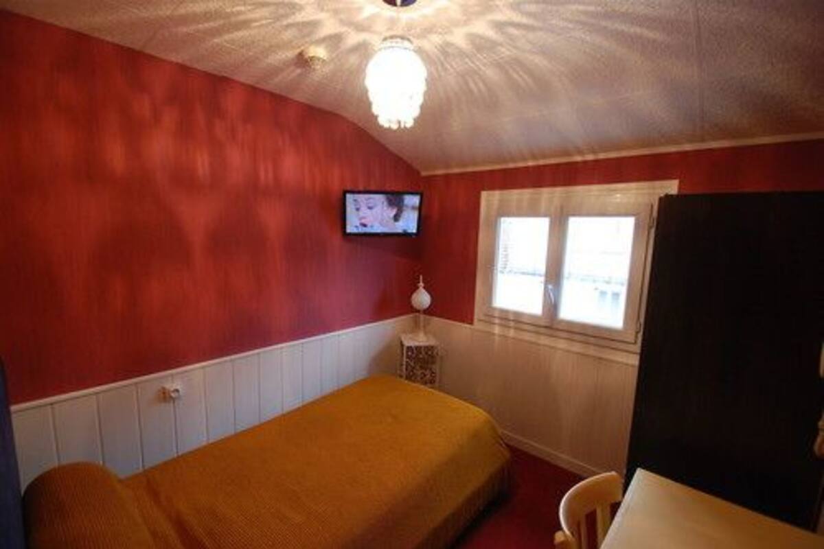 A small room with a single bed, a television mounted on the wall, and a window with blinds.
