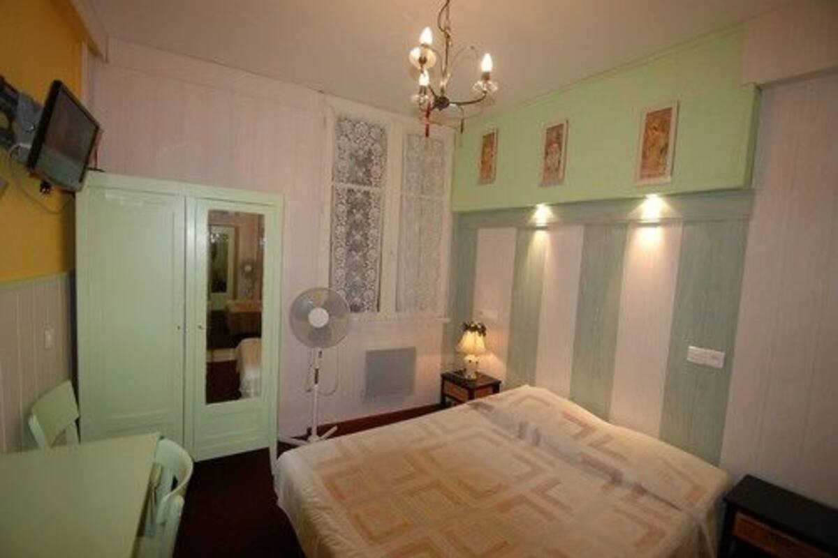 A room with a bed, a desk, a chair, a fan, a television, and a window with decorative shutters.