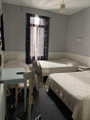 A hotel room with a desk, two beds, a chair, a standing fan, and a window with curtains. - Hotel Notre Dame Auxiliatrice (Lourdes)
