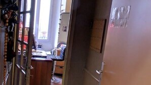 Un pasillo estrecho con una puerta que da a una habitación. El pasillo tiene piso de madera y una puerta con picaporte de metal. Hay una ventana con persianas en la puerta, y una pequeña señal en la pared.