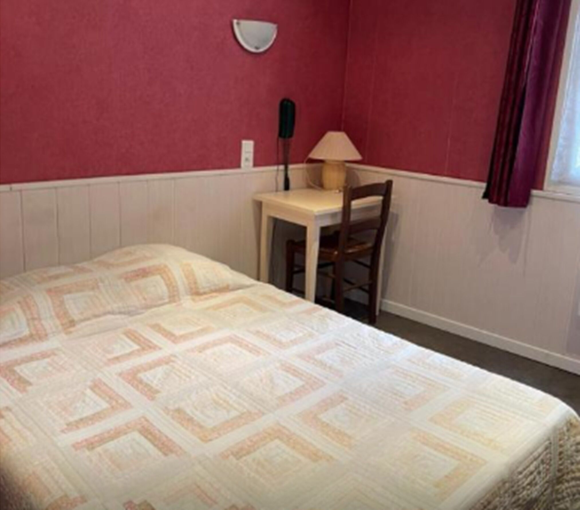 Una cama individual con una colcha estampada, una mesita con una lámpara y una silla en una habitación con paredes rojas y detalles blancos.
