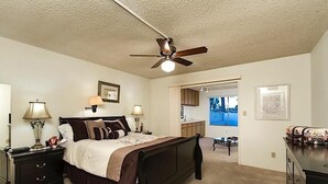 A bedroom with a bed, bedside tables, a ceiling fan, and a view of the kitchen and outside. - Sun Lakes By Signature Vacation Rentals (Chandler)