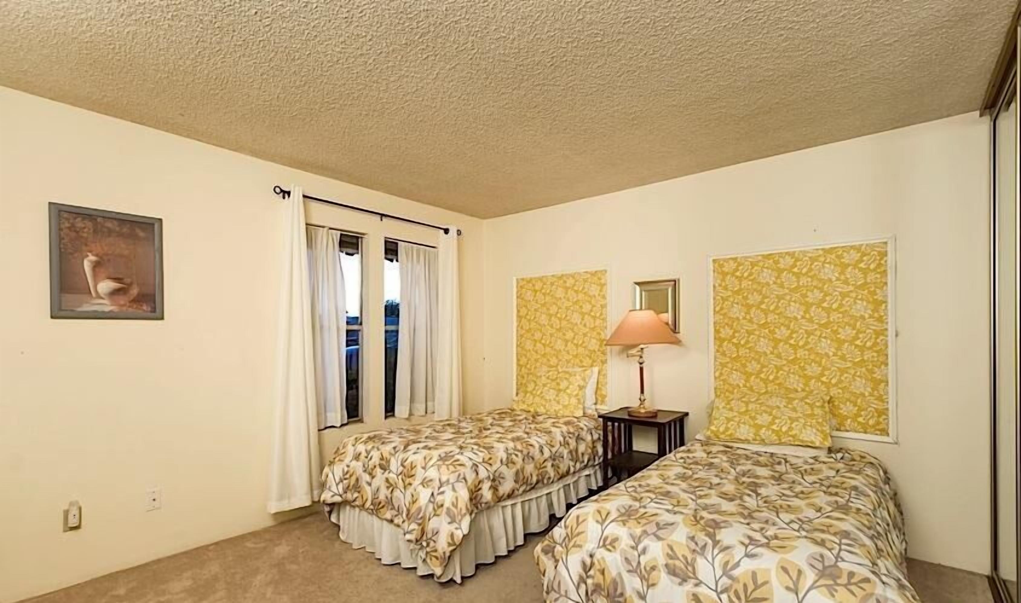 A bedroom with two single beds, a nightstand, a lamp, a window with curtains, and a wall-mounted mirror.
