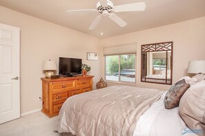 A bedroom with a bed, a TV on a wooden dresser, a ceiling fan, a window with a view, and a mirror. - Sweet Escape By Signature Vacation Rentals (Scottsdale)