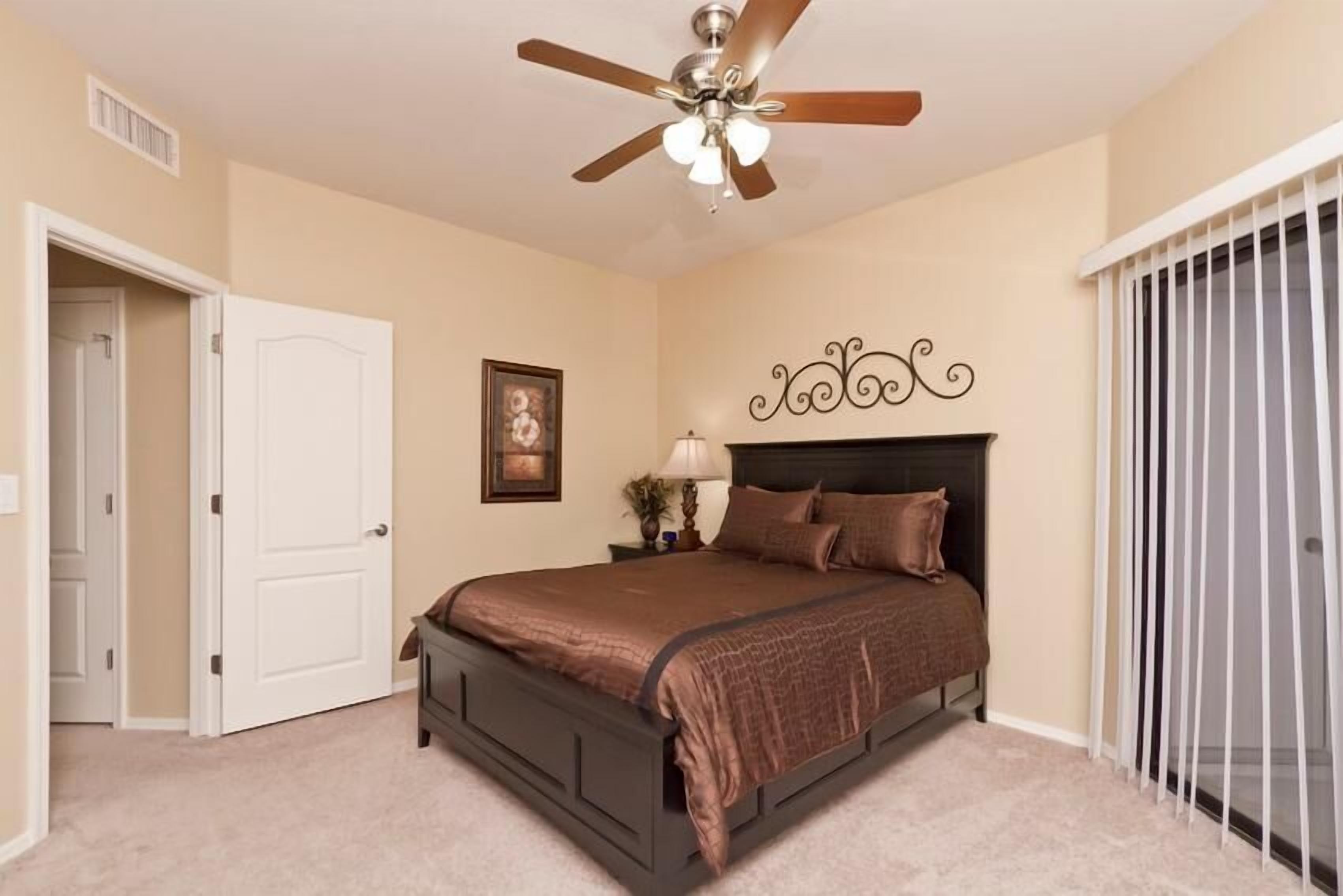 A bedroom with a bed, a ceiling fan, a door, a framed picture, and a window with blinds.
