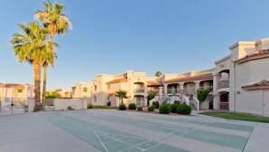 A courtyard with a shuffleboard court, palm trees, and a row of townhouses with balconies and a tiled roof. - Sunny Sky Condo By Signature Vacation Rentals (Peoria)