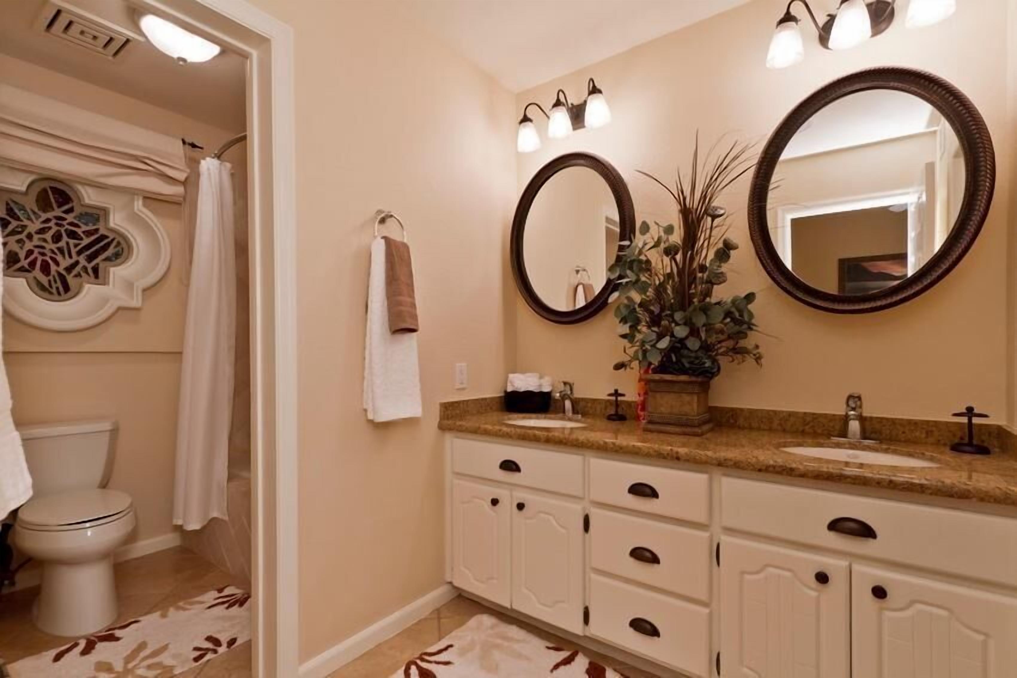 Une salle de bain comprenant des toilettes, un lavabo, un meuble-lavabo avec des placards, deux miroirs et un rideau de douche.