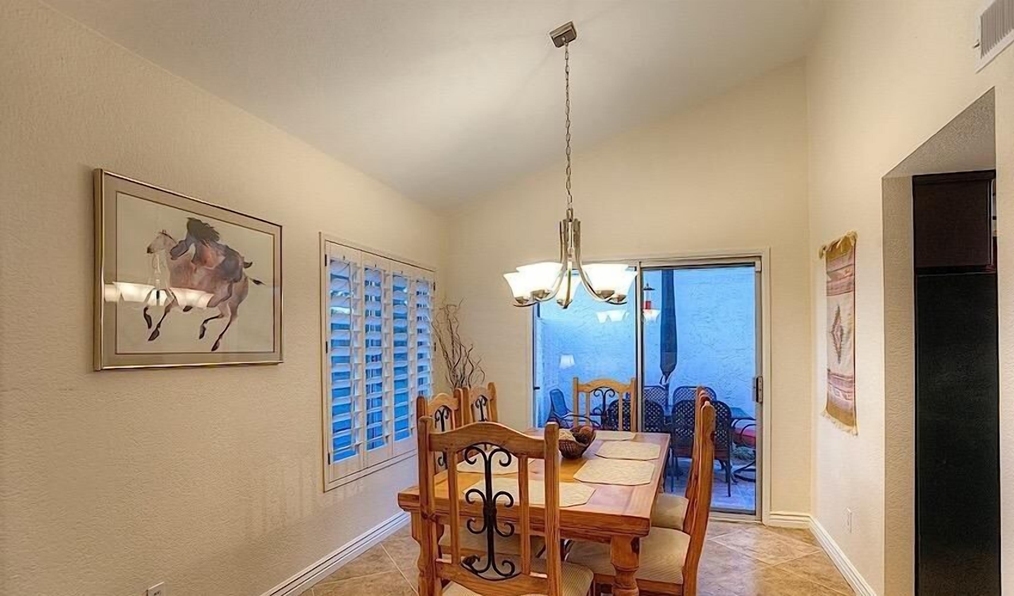 A dining area with a wooden table and chairs, a chandelier, a framed horse painting, and a window with blinds.