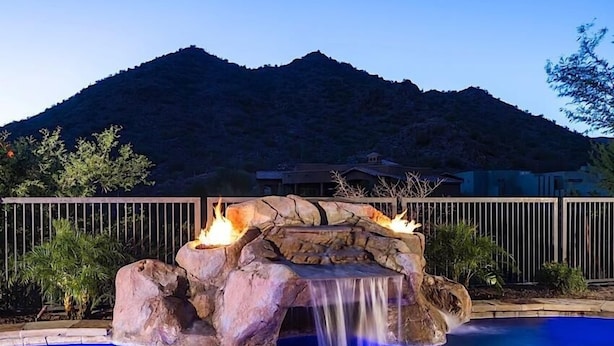 Una piscina con cascada de roca y un pozo de fuego, rodeada por una valla de madera y una montaña al fondo.