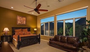A bedroom with a large bed, a ceiling fan, a window with a view of mountains, a brown leather couch, and a nightstand with a lamp. - South by Southwest - Signature Vacation Rentals (Scottsdale)