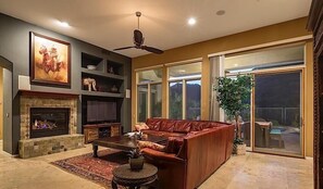 A spacious living room with a fireplace, leather sofa, and a large window overlooking a backyard. - South by Southwest - Signature Vacation Rentals (Scottsdale)