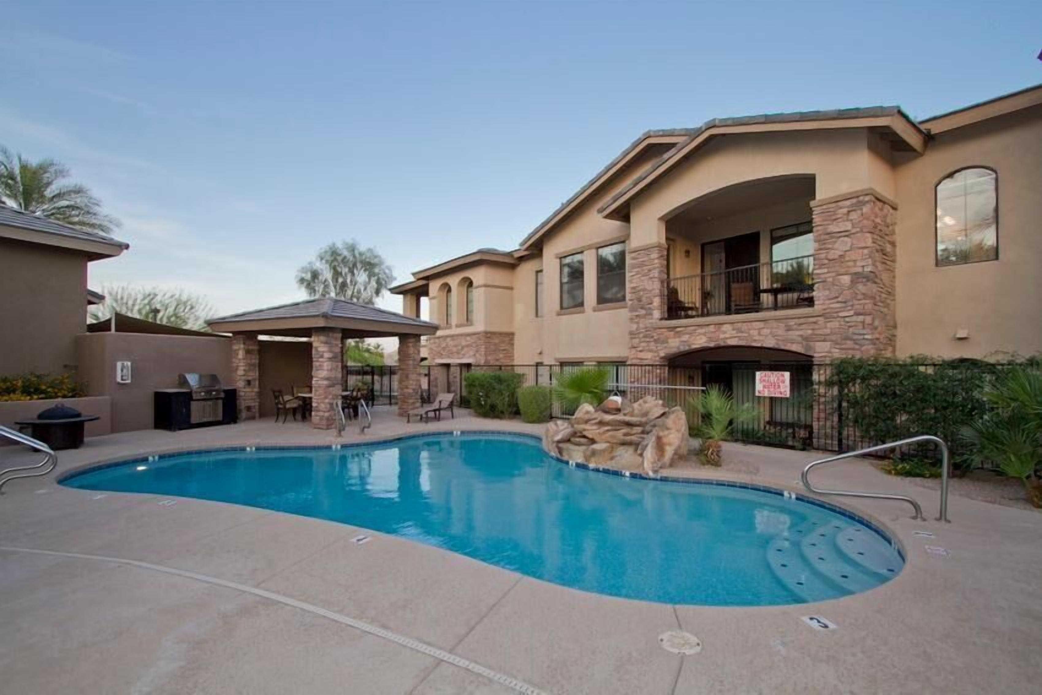 A swimming pool with a stone accent wall, a covered patio, and a two-story building with a balcony.