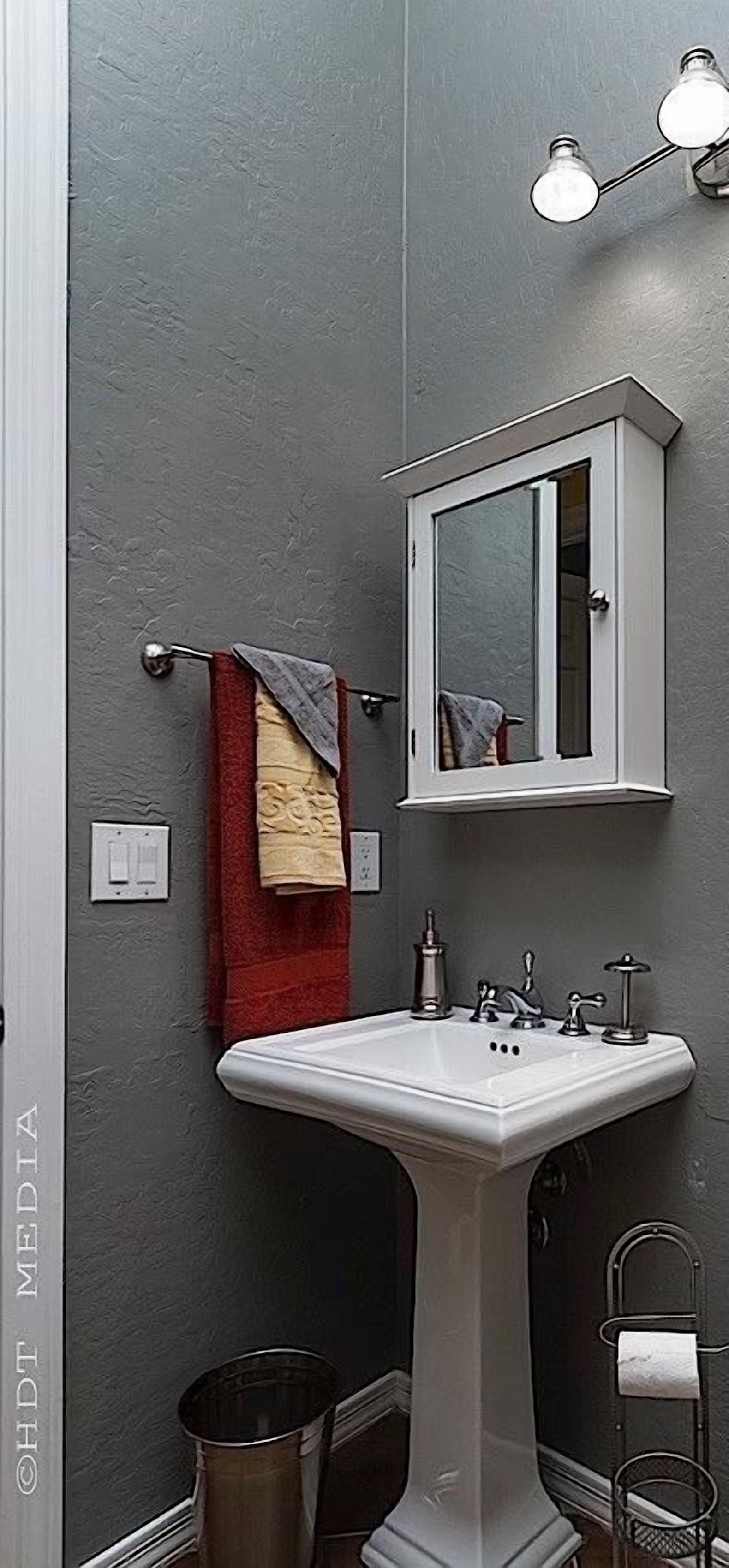 Une salle de bain avec un lavabo sur pied, un meuble à pharmacie blanc, un porte-serviettes avec une serviette rouge et un luminaire au-dessus du miroir.