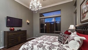 A bedroom with a large bed, a television, a chandelier, a window with a view, and a dresser.
