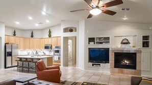 A modern living room with a fireplace, a television, and a kitchen area with wooden cabinets and a bar. - Sonoran Desert Retreat By Signature Vacation Rentals (Scottsdale)