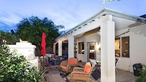 Un patio couvert, aménagé avec des meubles en osier et agrémenté d’un parasol rouge, le tout entouré de verdure.