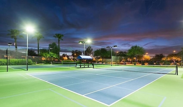 An outdoor tennis court at dusk with floodlights, surrounded by trees and a fence.