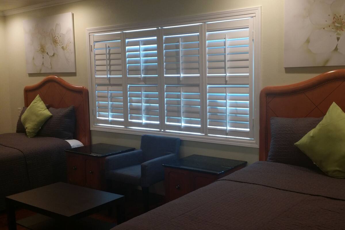 A hotel room with two beds, a small table, and a window with blinds.