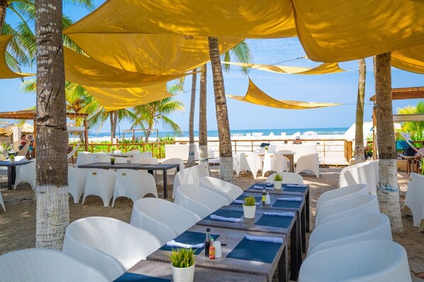 A beachside restaurant with outdoor seating, palm trees, and a view of the ocean.