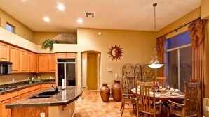 A kitchen with wooden cabinets, a granite countertop, a dining area with a table and chairs, and a sun decoration on the wall. - Desert Rose By Signature Vacation Rentals (Scottsdale)