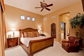 A bedroom with a wooden bed, two bedside lamps, a ceiling fan, a brown leather armchair, and a large plant. - Desert Rose By Signature Vacation Rentals (Scottsdale)