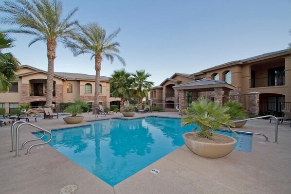 A swimming pool with lounge chairs and palm trees, surrounded by buildings with balconies and large windows.