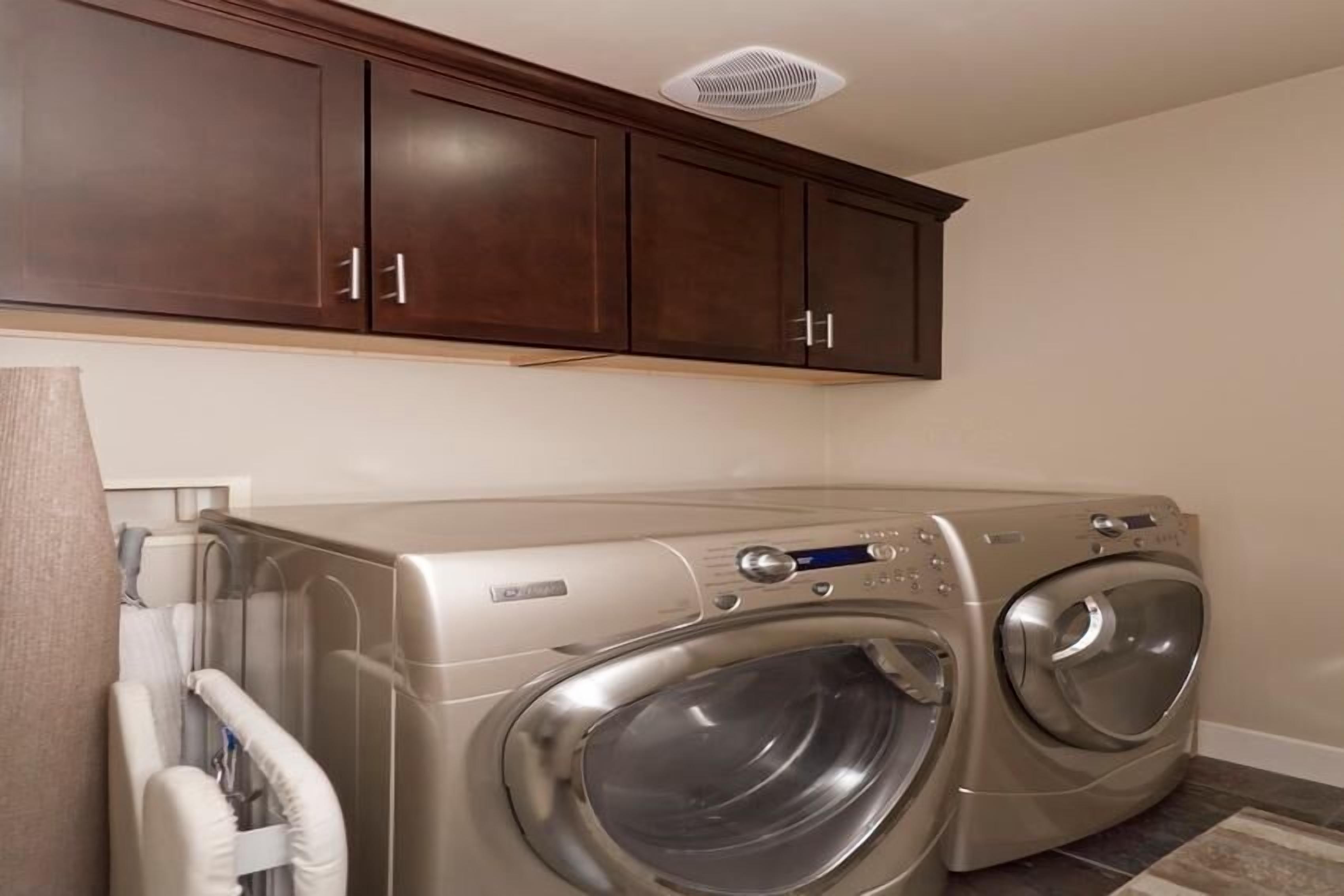 A laundry room with two front-loading washing machines, wooden cabinets above, and a vent on the ceiling.