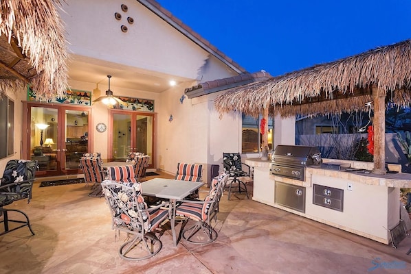 An outdoor patio with a dining table and chairs, a grill station, and a thatched roof.