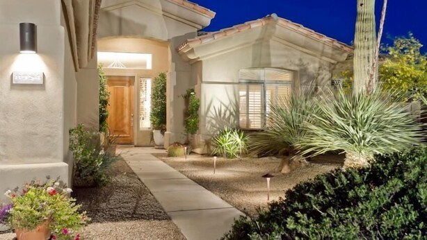 A well-lit residential property with a stone facade, a wooden door, and a landscaped garden featuring cacti and shrubs. - Casa De Luna By Signature Vacation Rentals (Scottsdale)