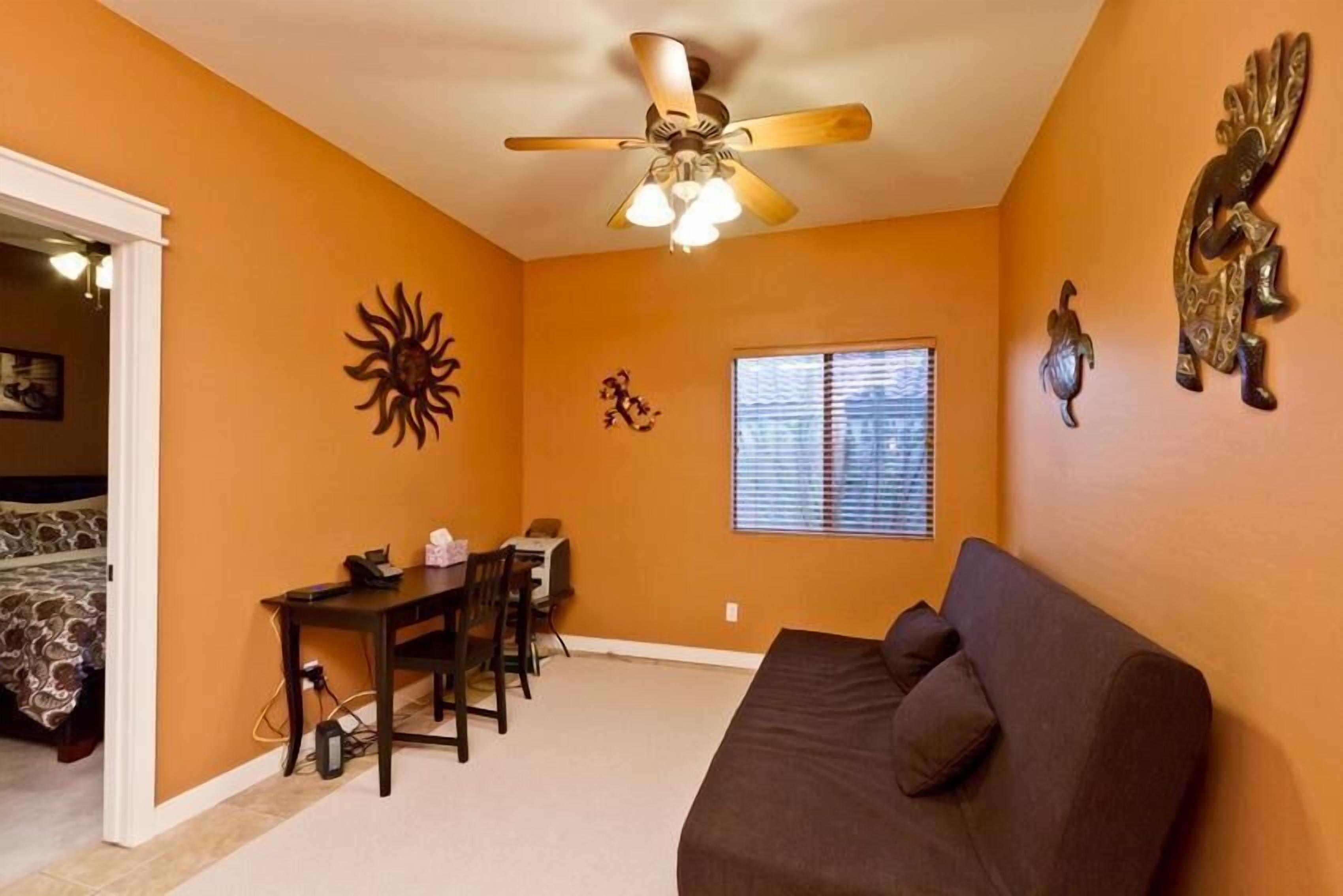 A room with orange walls, a ceiling fan, a window with blinds, a brown sofa, a dining table with chairs, and a bed with patterned bedding.