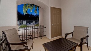 A balcony with two chairs and a table, a door, and a view of a fenced outdoor area with trees.