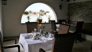 Un coin repas avec une table dressée pour quatre personnes, des chaises en osier, une nappe blanche et une vue à travers une fenêtre en arc de cercle.