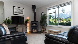 A living room with a fireplace, a television, and a large window with a mountain view. - The Cabin at Sweetwater Lane Farm (Mount Currie)