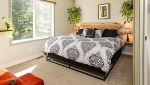 A bedroom with a large bed, two bedside lamps, a wooden headboard, a window with a view of trees, and two hanging plants. - The Cabin at Sweetwater Lane Farm (Mount Currie)
