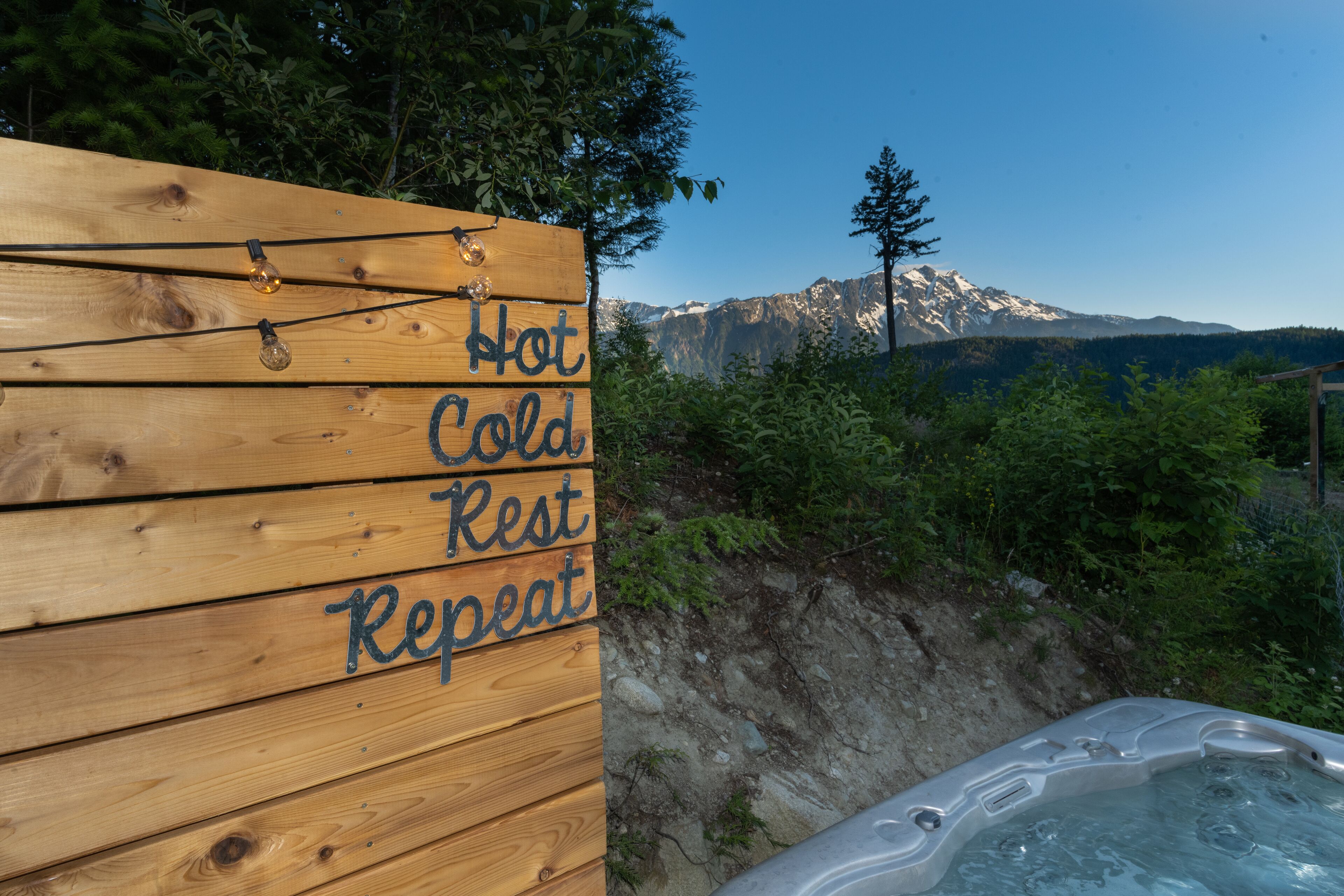 A wooden sign with the words "Hot Cold Rest Repeat" and a hot tub with a mountainous backdrop.