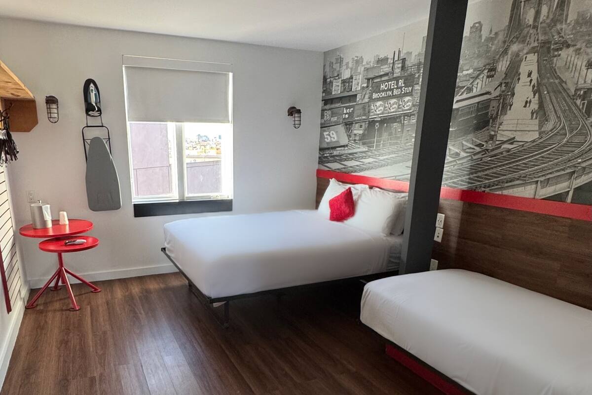 Camera d'albergo compatta con due letti, un piccolo tavolo rosso e una finestra vista città.