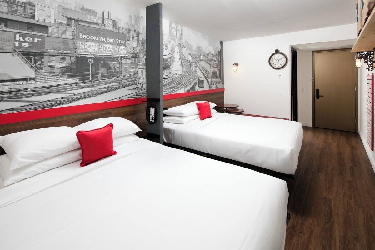 Camera d'albergo con due letti, una parete decorata in rosso, un grande murale in bianco e nero raffigurante una città e un pavimento in legno.
