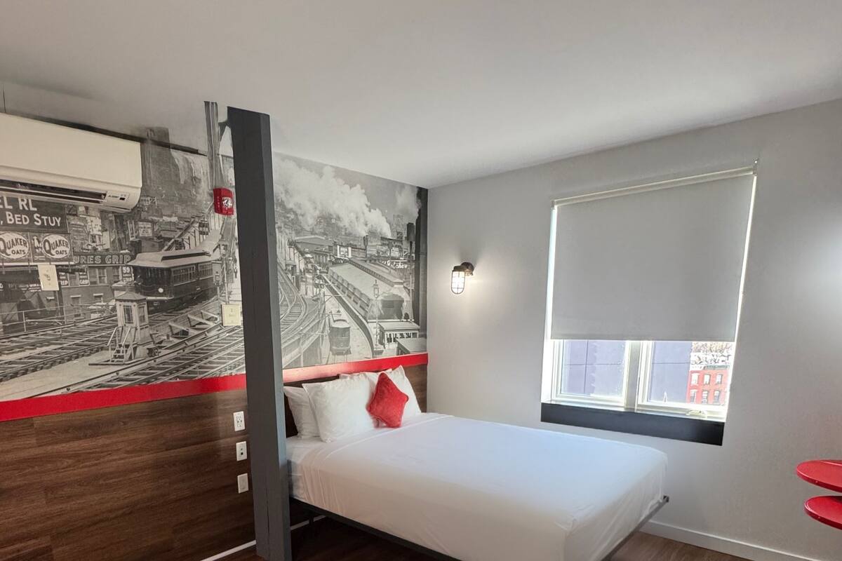 Una camera d'albergo moderna con un letto, un murale raffigurante un panorama urbano, una finestra con vista e una sedia rossa.
