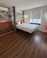 Quarto de hotel moderno com uma cama, um mural com vista para a cidade, uma janela e uma cadeira vermelha.
