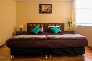 A double bed with a wooden headboard, two teal pillows, and a dark brown bedspread in a room with wooden flooring and a window on the right.